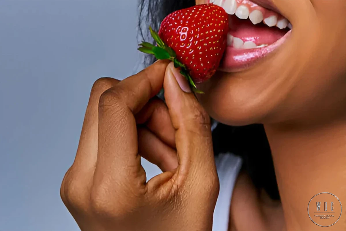 Kobieta gryząca kawałek truskawki i uśmiechająca się pięknym uśmiechem.