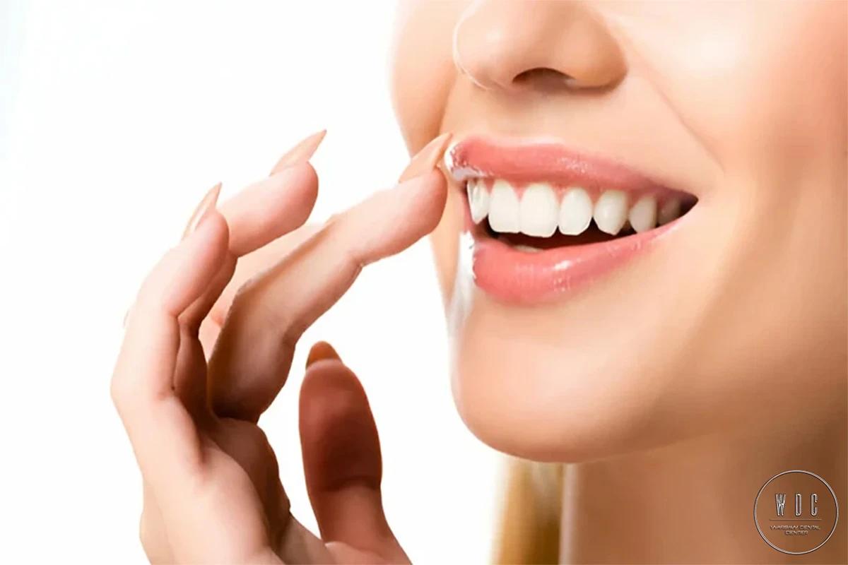 Kobieta uśmiecha się i dotyka ust, pokazując swoje proste, piękne zęby.
